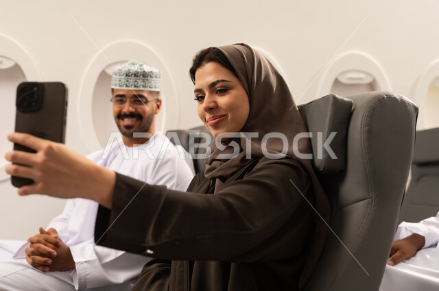 مفهوم الزواج والارتباط, امراة عربيه خليجيه عمانية ترتدي الحجاب و العباءة تجلس داهل الطائرة فى رحله جوية برفقة زوجها تقوم بالتقاط صور السيلفي التذكارية باستخدام الهاتف المحمول، قضاء أوقات ودية بين الزوجين، توثيق اللحظات السعيدة، أجهزة وتقنيات حديثة