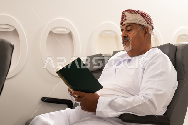 The love of reading and literature, spending free time learning and educating oneself, the interest in browsing useful books, an elderly Arab Gulf Omani man wearing a dishdasha and kumma sitting inside an airplane on a flight, looking at the book with contemplation and focus, the culture of self-education, the concept of tourism and travel.