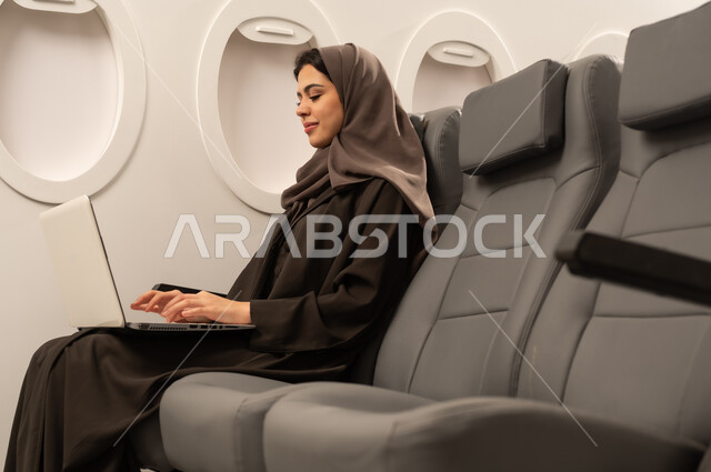 إنجاز المهام المطلوبة عبر اللابتوب, ادارة الأعمال عن بعد، استخدام الأجهزة والتقنيات الحديثة والمتطورة، إمرأة عربية خليجية عمانية ترتدي العباءة والحجاب تجلس داخل الطائرة فى رحلة جوية تقوم باستخدام الحاسب المحمول، ايماءات الاندماج و التركيز، دمج التكنولوجيا بالعمل, مفهوم السياحة و السفر