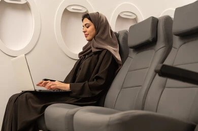 إنجاز المهام المطلوبة عبر اللابتوب, ادارة الأعمال عن بعد، استخدام الأجهزة والتقنيات الحديثة والمتطورة، إمرأة عربية خليجية عمانية ترتدي العباءة والحجاب تجلس داخل الطائرة فى رحلة جوية تقوم باستخدام الحاسب المحمول، ايماءات الاندماج و التركيز، دمج التكنولوجيا بالعمل, مفهوم السياحة و السفر
