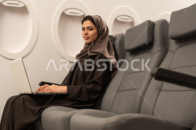 دمج التكنولوجيا بالعمل, متابعة الأعمال والمهام عن بعد، استخدام الأجهزة والتقنيات الحديثة والمتطورة، إمرأة عربية خليجية عمانية ترتدي العباءة والحجاب تجلس داخل الطائرة فى رحلة جوية تقوم باستخدام الحاسب المحمول، ايماءات السعادة و السرور، إنجاز المهام المطلوبة عبر اللابتوب, مفهوم السياحة و السفر