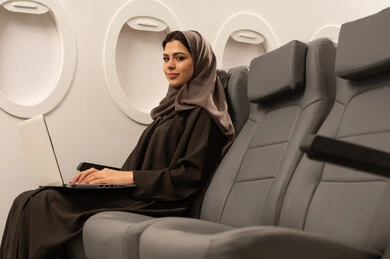 دمج التكنولوجيا بالعمل, متابعة الأعمال والمهام عن بعد، استخدام الأجهزة والتقنيات الحديثة والمتطورة، إمرأة عربية خليجية عمانية ترتدي العباءة والحجاب تجلس داخل الطائرة فى رحلة جوية تقوم باستخدام الحاسب المحمول، ايماءات السعادة و السرور، إنجاز المهام المطلوبة عبر اللابتوب, مفهوم السياحة و السفر