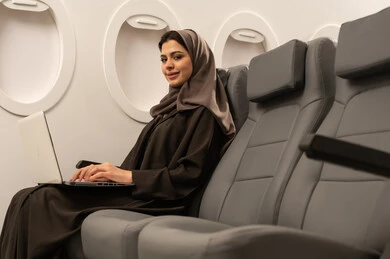 Supervising work remotely, using modern and advanced devices and technologies, an Arab Gulf Omani woman wearing an abaya and hijab sitting inside an airplane on a flight using a laptop, gestures of happiness and joy, integrating technology into work, completing required tasks via the laptop, the concept of tourism and travel.