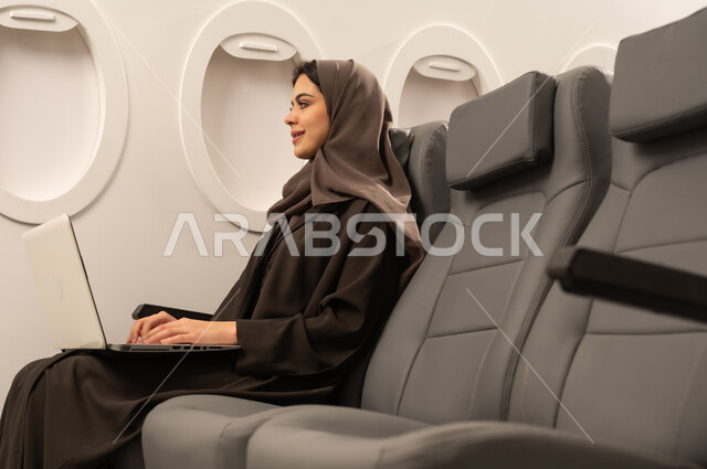 ادارة الأعمال عن بعد، استخدام الأجهزة والتقنيات الحديثة والمتطورة، إمرأة عربية خليجية عمانية ترتدي العباءة والحجاب تجلس داخل الطائرة فى رحلة جوية تقوم باستخدام الحاسب المحمول، ايماءات السعادة و السرور، دمج التكنولوجيا بالعمل، إنجاز المهام المطلوبة عبر اللابتوب, مفهوم السياحة و السفر