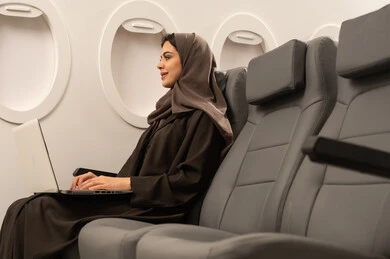 Remote business management, using modern and advanced devices and technologies, an Arab Gulf Omani woman wearing an abaya and hijab sitting inside an airplane on a flight using a laptop, gestures of happiness and joy, integrating technology with work, completing required tasks via the laptop, the concept of tourism and travel.
