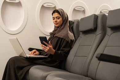 النظر إلى الجوال ومتابعة آخر المستجدات, تصفح مواقع وصفحات الانترنت عبر الهاتف المحمول, التواصل مع العائلة والأصدقاء, امراة عربيه خليجيه عمانية ترتدي الحجاب و العباءة تجلس داخل الطائرة فى رحلة جويه تعمل على الحاسب المحمول, مفهوم السياحة و السفر