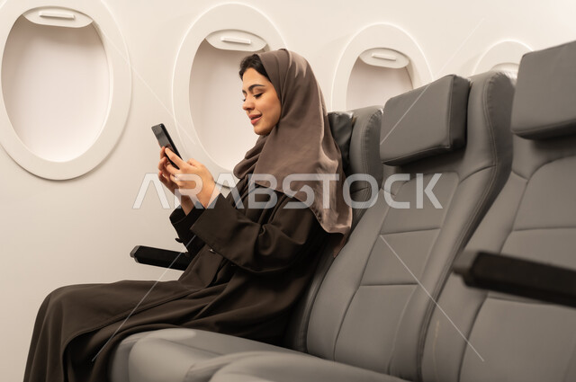 Browsing social media sites, communicating through modern electronic applications, a side view of a smiling Omani Gulf Arab woman wearing a hijab sitting inside an airplane during a flight, talking to friends via mobile phone, the concept of tourism and travel.