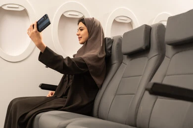 Taking a commemorative photo, chatting with family and friends through the mobile via video call, using modern advanced devices and technologies, an Arab Gulf Omani woman wearing a hijab and a traditional abaya sits inside the airplane on a flight, the concept of tourism and travel.