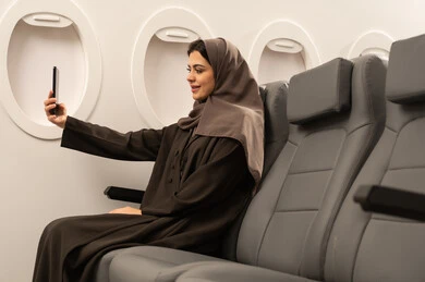 إجراء مكالمة فيديو مع العائلة والأصدقاء عبر الهاتف, التقاط صورة للذكرى , استخدام الاجهزة والتقنيات الحديثة المتطورة، امراة عربية هليجيه عمانية ترتدي الحجاب و العباءة الدشداشة و الكم تجلس داخل الطائرة فى رحلة جوية ، مفهوم السياحة و السفر
