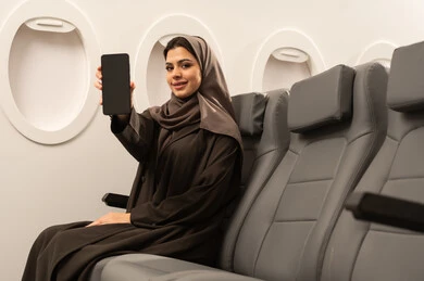 The use of advanced modern devices and technologies, displaying a blank black screen on a mobile phone, an Arab Gulf Omani woman wearing a hijab and abaya sitting inside an airplane on a flight, the concept of tourism and travel, gestures of happiness and joy.