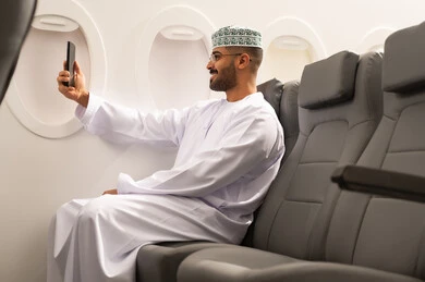 التقاط صورة شخصية تذكارية, الدردشة مع الاهل و الاصدقاء من خلال الجوال عبر مكالمة فيديو , استخدام الاجهزة والتقنيات الحديثة المتطورة، رجل عربي خليجي عماني يرتدي الدشداشة و الكمه يجلس داخل الطائرة فى رحلة جوية ، مفهوم السياحة و السفر
