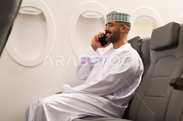 Chatting with family and friends through mobile, using modern advanced devices and technologies, an Arab Gulf Omani man wearing a dishdasha and kumma sitting inside an airplane on a flight, the concept of tourism and travel.