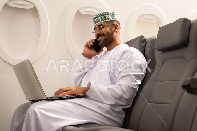 اجراء مكالمه هاتفية عبر الجوال, التواصل مع الأهل والاصدقاء عبر اللابتوب، انجاز مهام العمل عن بعد بايماءات الاندماج، رجل عربي خليجي عماني يرتدي االدشداشة و الكمة يجلس في الطائرة ويعمل على الحاسب المحمول، استخدام الأجهزة والتقنيات الحديثة، مفهوم السياحة والسفر