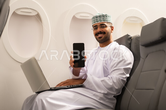 عرض شاشة فارغة سوداء عبر الجوال , التواصل مع الأهل والاصدقاء عبر اللابتوب، انجاز مهام العمل عن بعد بايماءات الاندماج، رجل عربي خليجي عماني يرتدي االدشداشة و الكمة يجلس في الطائرة ويعمل على الحاسب المحمول، استخدام الأجهزة والتقنيات الحديثة، مفهوم السياحة والسفر