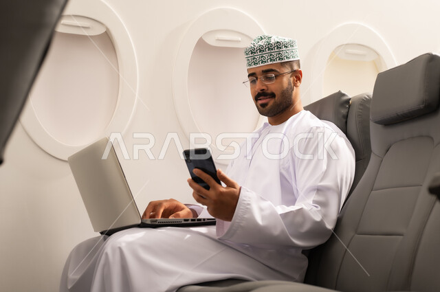 Browsing social media, completing remote work tasks with integration gestures, an Arab Gulf Omani man wearing a dishdasha and kumma sitting on a plane using a laptop and mobile phone, using modern devices and technologies, the concept of tourism and travel.