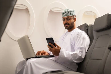 تصفح مواقع التواصل الالكتروني، انجاز مهام العمل عن بعد بايماءات الاندماج، رجل عربي خليجي عماني يرتدي االدشداشة و الكمة يجلس في الطائرة يستخدم الحاسب المحمول و الجوال ، استخدام الأجهزة والتقنيات الحديثة، مفهوم السياحة والسفر
