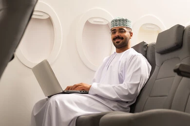 النظر الى الكاميرا بتعابير السعاده , التواصل مع الأهل والاصدقاء عبر اللابتوب، انجاز مهام العمل عن بعد بايماءات الاندماج، رجل عربي خليجي عماني يرتدي االدشداشة و الكمة  يجلس في الطائرة ويعمل على الحاسب المحمول، استخدام الأجهزة والتقنيات الحديثة، مفهوم السياحة والسفر