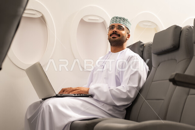 النظر الى شيء ما , التواصل مع الأهل والاصدقاء عبر اللابتوب، انجاز مهام العمل عن بعد بايماءات الاندماج، رجل عربي خليجي عماني يرتدي االدشداشة و الكمة  يجلس في الطائرة ويعمل على الحاسب المحمول، استخدام الأجهزة والتقنيات الحديثة، مفهوم السياحة والسفر