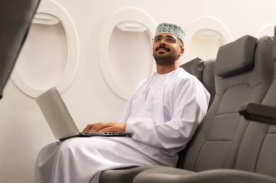 Looking at something, communicating with family and friends via laptop, completing work tasks remotely with integration gestures, an Arab Gulf Omani man wearing a dishdasha and kumma sitting on a plane and working on a laptop, using modern devices and technologies, the concept of tourism and travel.