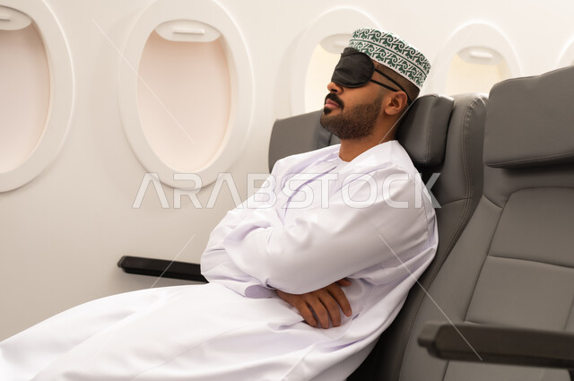 وسائل مواصلات جوية, ايماءات تدل على النوم والاسترخاء ، رجل عربي خليجي عماني يرتدي الدشداشة و الكمة يضع قناع النوم على عينيه يجلس في مقصورة الطائرة ، الشعور بالتعب والارهاق  ، خطوط الطيران الجوي ، سياحة وسفر ، رحلات جوية سياحية