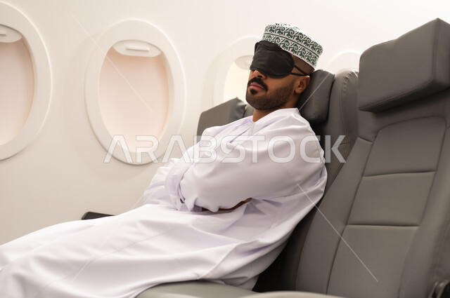 Feeling tired and exhausted, gestures indicating sleep and relaxation, an Arab Gulf Omani man wearing a dishdasha and kumma puts a sleep mask over his eyes sitting in the airplane cabin, air transportation means, airline companies, tourism and travel, tourist flights.