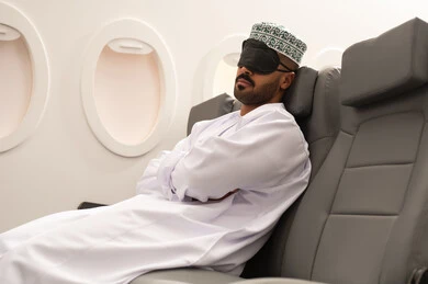 الشعور بالتعب والارهاق, ايماءات تدل على النوم والاسترخاء ، رجل عربي خليجي عماني يرتدي الدشداشة و الكمة  يضع قناع النوم على عينية يجلس في مقصورة الطائرة  ، وسائل مواصلات جوية ، خطوط الطيران الجوي ، سياحة وسفر ، رحلات جوية سياحية
