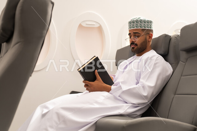 مفهوم الشغف بالقراءة, قراءة الكتب المفيدة للتعلم والتثقف, خطوط الطيران العماني, قضاء أوقات ممتعه في مقصورة الطائرة, رجل عربي خليجي عماني يرتدي الدشداشة و الكمه يجلس فى مقعد الطائرة,  الاستعداد قضاء عطله صيفية