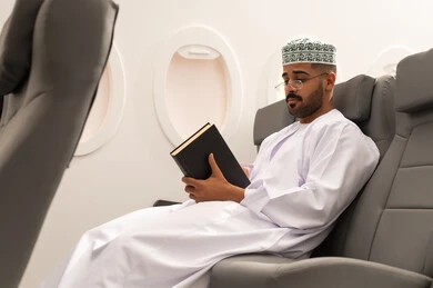 مفهوم الشغف بالقراءة, قراءة الكتب المفيدة للتعلم والتثقف, خطوط الطيران العماني, قضاء أوقات ممتعه في مقصورة الطائرة, رجل عربي خليجي عماني يرتدي الدشداشة و الكمه يجلس فى مقعد الطائرة,  الاستعداد قضاء عطله صيفية