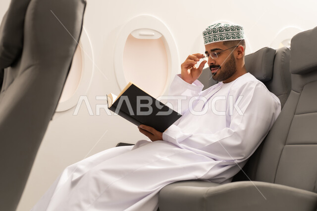 Reading useful books for learning and culture, Oman Air, spending enjoyable times in the airplane cabin, practicing the hobby of reading, an Arab Gulf Omani man wearing a dishdasha and kumma sitting in an airplane seat, preparing to spend a summer vacation.