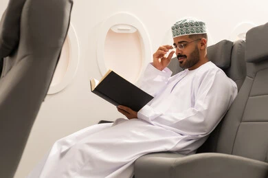 قراءة الكتب المفيدة للتعلم والتثقف, خطوط الطيران العماني, قضاء أوقات ممتعه في مقصورة الطائرة, ممارسة هواية القراءه, رجل عربي خليجي عماني يرتدي الدشداشة و الكمه يجلس فى مقعد الطائرة,  الاستعداد لقضاء عطله صيفية