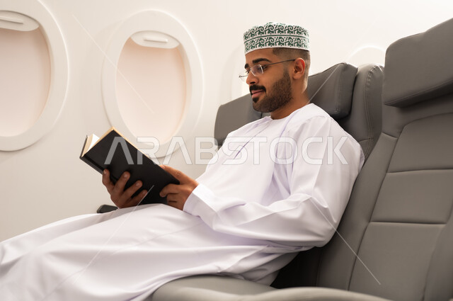 ممارسة هواية القراءه, خطوط الطيران العماني, قضاء أوقات ممتعه في مقصورة الطائرة, قراءة الكتب المفيدة للتعلم والتثقف, رجل عربي خليجي عماني يرتدي الدشداشة و الكمه يجلس فى مقعد الطائرة,  الاستعداد لقضاء عطله صيفية