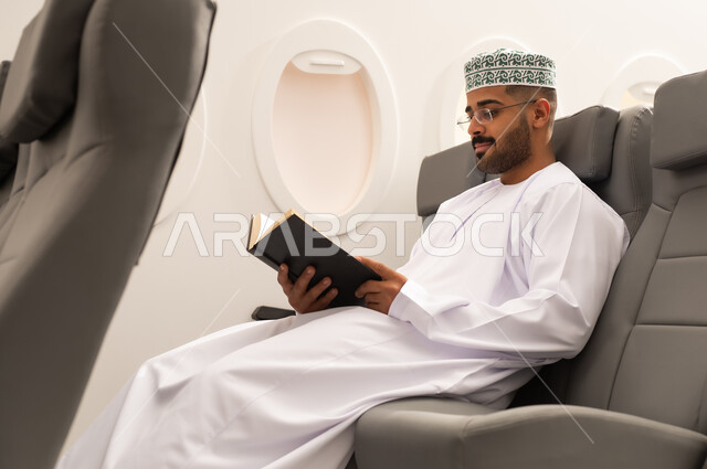 خطوط الطيران العماني, قضاء أوقات ممتعه في مقصورة الطائرة, ممارسة هواية القراءه, قراءة الكتب المفيدة للتعلم والتثقف, رجل عربي خليجي عماني يرتدي الدشداشة و الكمه يجلس فى مقعد الطائرة,  الاستعداد قضاء عطله صيفية