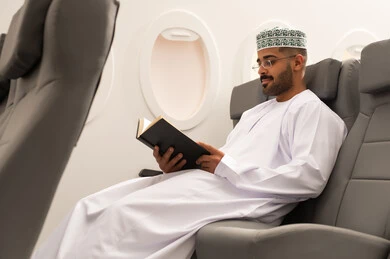 Oman Air, spending enjoyable times in the airplane cabin, practicing the hobby of reading, reading useful books for learning and culture, an Arab Gulf Omani man wearing a dishdasha and kumma sitting in an airplane seat, preparing to spend a summer vacation.