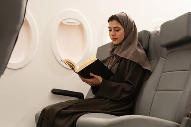 Practicing the hobby of reading, spending enjoyable times in the airplane cabin, reading useful books for learning and culture, an Arab Khaleeji Omani woman wearing a hijab and abaya sitting in an airplane seat, spending a summer vacation, Oman Air.
