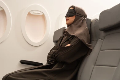 رحلات جوية سياحية, الشعور بالارهاق والتعب اثناء السفر, امراة عربية خليجية عمانية محجبة ترتدي العباءة تضع قناع النوم على عينيها تجلس داخل مقصورة الطائرة ، ايماءات تدل على النوم والاسترخاء ، وسائل مواصلات جوية ، خطوط الطيران الجوي ، سياحة وسفر