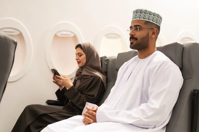 استخدام تطبيقات الهاتف المحمول، التحدث أونلاين مع الأصدقاء، زوجان عربيان خليجيان عمانيان يرتديان الزي التقليدي يجلسان داخل الطائرة فى رحله جوية امراة خليجية تقوم بمتابعة اخبار وسائل التواصل الاجتماعي باستخدام الجوال، ايماءات الاندماج والتركيز, مفهوم السياحة و السفر