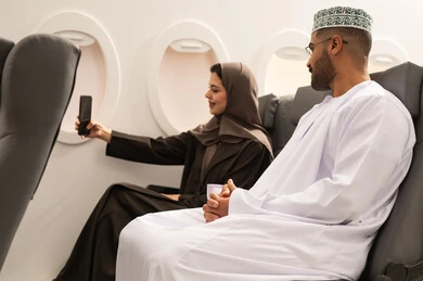 التقاط صورة شخصية تذكاريةعبر الهاتف المحمول , استخدام جهاز تقني حديث, دمج التكنولوجيا المتطورة بالحياة اليومية، امراة عربيه خليجيه عمانية ترتدي الحجاب و العباءة تجلس داخل الطائرة فى رحلة جوية برفقة زوجها تقوم بأخذ لقطة سيلفي , ايماءات السعاده و السرور