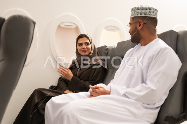 الدردشات والحوارات المتبادلة, قضاء وقت ممتع خلال فترة السفر, زوجان عربيان خليجيان عمانيان يرتديان الزي التقليدي يجلسان داخل الطائرة فى رحله جوية, مفهوم السياحه و السفر