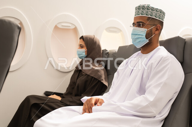 قضاء وقت ممتع خلال فترة السفر, زوجان عربيان خليجيان عمانيان يرتديان الزي التقليدي  و قناع الحماية يجلسان داخل الطائرة فى رحله جوية, توخى الحذر من انتشار فيروس كورونا , مفهوم السياحة و السفر