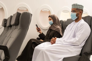 Spending enjoyable times while commuting and traveling, an Arab Gulf couple from Oman wearing traditional attire and a coronavirus protection mask sitting inside the airplane on a flight, a Gulf woman holding a mobile phone in her hands, the concept of tourism and travel.