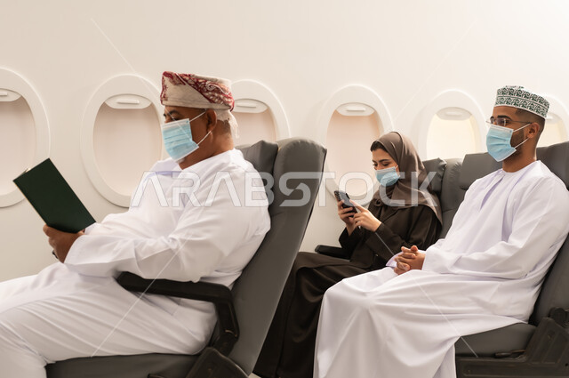 حب المطالعة والقراءة, الاهتمام بتصفح الكتب المفيدة، رجل عربي خليجي عماني مسن يرتدي الدشداشه والكمه و قناع كورونا يجلس داخل الطائرة فى رحله جوية ينظر الى الكتاب بتمعن وتركيز, امراة عربيه خليجيه عمانية ترتدي العباءة تستخدم الهاتف المحمول