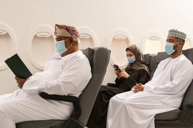 The love of reading and literature, the interest in browsing useful books, an elderly Arab Gulf Omani man wearing a dishdasha and a mask sits inside the airplane on a flight, looking at the book with contemplation and focus, an Arab Gulf Omani woman wearing an abaya is using a mobile phone.