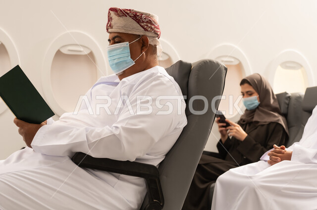الاهتمام بتصفح الكتب المفيدة، حب المطالعة والقراءة، رجل عربي خليجي عماني مسن يرتدي الدشداشه والكمه و قناع كورونا يجلس داخل الطائرة فى رحله جوية ينظر الى الكتاب بتمعن وتركيز, امراة عربيه خليجيه عمانية ترتدي العباءة تستخدم الهاتف المحمول