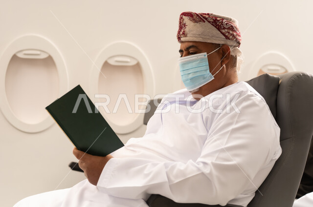 قضاء وقت الفراغ بالتعلم والتثقف، الاهتمام بتصفح الكتب المفيدة، حب المطالعة والقراءة، رجل عربي خليجي عماني مسن  يرتدي الدشداشه والكمه و قناع كورونا يجلس داخل الطائرة فى رحله جوية ينظر الى الكتاب بتمعن وتركيز، ثقافة التعليم الذاتي, مفهوم السياحة و السفر