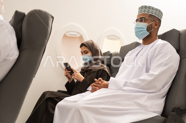 الاستمتاع بوقت الفراغ اثناء السفر, رجل عربي خليجي يرتدي الدشداشة و الكمة و قناع كورونا يجلس داخل الطائرة اثناء رحله جوية برفقه زوجته , امراة خليجيه تمسك بيديها الهاتف المحمول , مفهوم السياحة و السفر