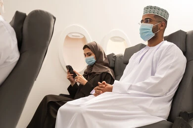 الاستمتاع بوقت الفراغ اثناء السفر, رجل عربي خليجي يرتدي الدشداشة و الكمة و قناع كورونا يجلس داخل الطائرة اثناء رحله جوية برفقه زوجته , امراة خليجيه تمسك بيديها الهاتف المحمول , مفهوم السياحة و السفر