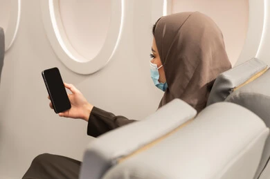 Talking to relatives and family via video call, following the latest news on social media, a close-up shot from behind of an Emirati Gulf Arab woman wearing a hijab and abaya and a coronavirus protection mask sitting inside an airplane on a flight browsing social media, the concept of tourism and travel.