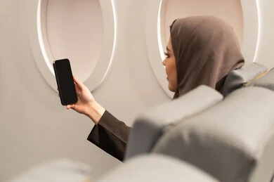 Making a video call with family and relatives via mobile, following the latest news on social media, a close-up shot from behind of an Arab Gulf Emirati woman wearing a hijab and abaya sitting inside the airplane on a flight browsing social media, the concept of tourism and travel.