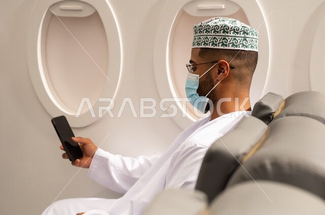 دمج التكنولوجيا المتطورة بالحياة اليومية, الدردشة مع الأهل والأصدقاء، تصفح مواقع التواصل الاجتماعي، استخدام جهاز تقني حديث، صورة من الجانب لرجل عربي خليجي عماني يرتدي الدشداشة والكمة يرتدي قناع حماية كورونا يجلس داخل الطائرة فى رحله جوية يستخدم الهاتف المحمول, مفهوم السياحة و السفر