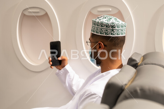 استخدام جهاز تقني حديث, الدردشة مع الأهل والأصدقاء، تصفح مواقع التواصل الاجتماعي, دمج التكنولوجيا المتطورة بالحياة اليومية، صورة من الجانب لرجل عربي خليجي عماني يرتدي الدشداشة والكمة يرتدي قناع حماية كورونا يجلس داخل الطائرة فى رحله جوية يستخدم الهاتف المحمول, مفهوم السياحة و السفر